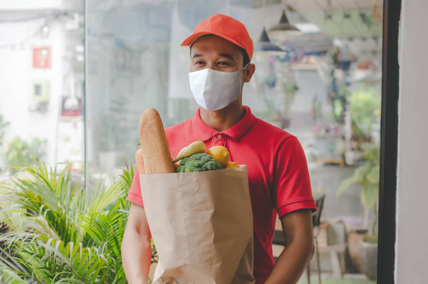 food delivery service man with protection face mask in red uniform holding fresh food set bag waiting for customer at door home, healthy food, express delivery, food delivery, online shopping concept - 写真・画像