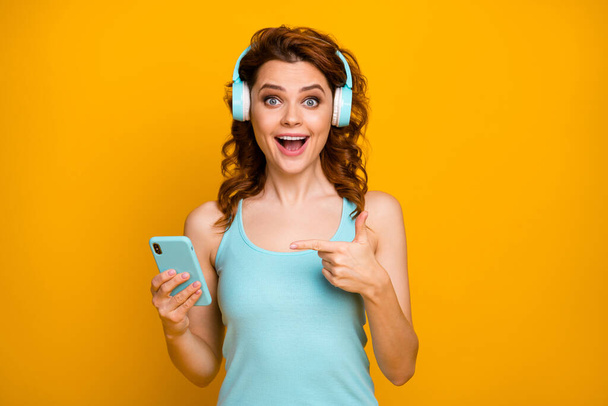 I love this song. Photo of pretty funny lady open mouth direct finger telephone listen headphones audio good mood wear casual teal tank-top isolated vibrant yellow color background - Photo, Image
