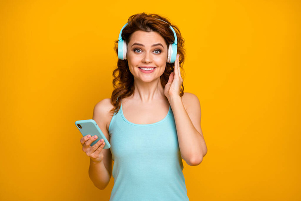 Photo of pretty funny lady hold telephone listen modern technology headphones popular songs good mood wear casual teal tank-top isolated vibrant yellow color background - Foto, immagini