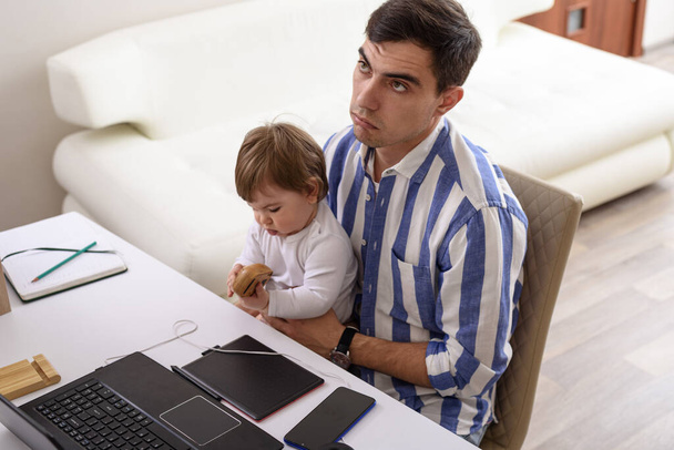 tired dad with baby on his lap sitting at laptop, fatherhood, dad on maternity leave, remote work at home concept - Photo, Image