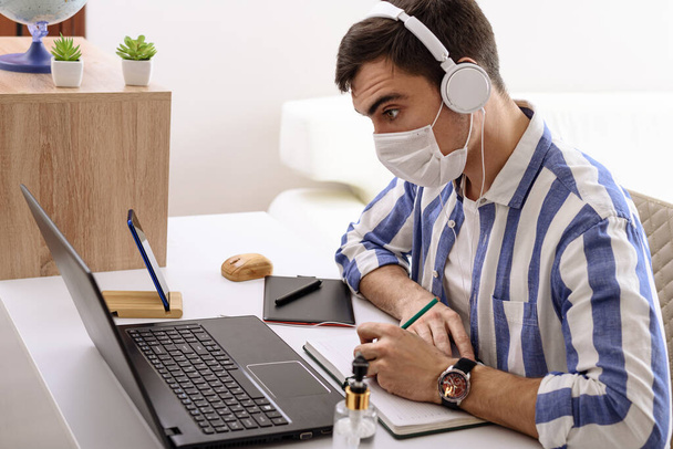 caucasian man in mask and headphones listens to music, writes in notebook, remote work at home, safe work, freelancer, self-isolation - Photo, Image