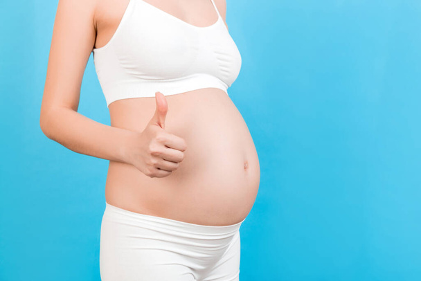 Beyaz iç çamaşırlı, pozitif hamile bir kadının mavi arka plandaki bebek şişkinliğinin üzerinde başparmak hareketi yaparken çekilmiş görüntüler. Doğum günün kutlu olsun. Boşluğu kopyala. - Fotoğraf, Görsel