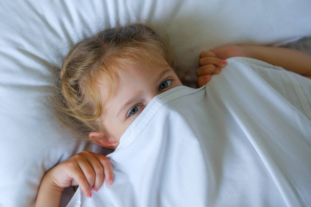 Güzel küçük kız sabah uyanır ve battaniyenin altında yatar. Çocuk okula uyandı.. - Fotoğraf, Görsel