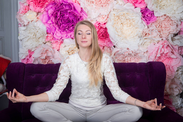 krásná blondýna dívka v ležérní oblečení sedí na fialové staromódní pohovka v jóga lotos pozice na velké růžové pivoňky pozadí - Fotografie, Obrázek
