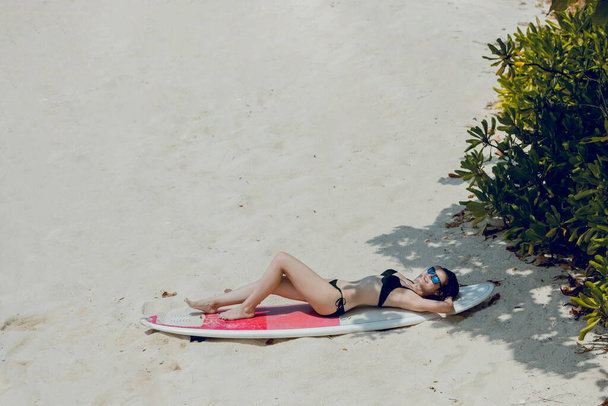 Belle jeune femme brune au corps mince allongé sur la plage avec planche de surf blanche et rouge pendant les vacances actives dans l'île tropicale. Style de vie de surf et concept d'aventure de sport nautique extrême
. - Photo, image