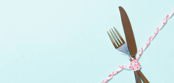 Dieet en gezond eten concept met gekruiste vork en mes en gekrulde meetlint op blauwe achtergrond. Bovenaanzicht van gewichtsverlies met kopieerruimte. - Foto, afbeelding