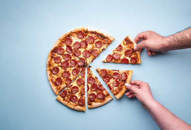 Mann und Frau greifen nach Pizzascheiben. Aufgeschnittene Paprika-Pizza auf blauem Hintergrund, über der Ansicht. Draufsicht mit leckerer Pfefferoni-Pizza. - Foto, Bild