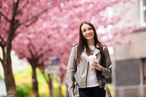 brunette girl in jacket and jeans in the spring outdoors in the city against the backdrop of flowering sakura trees posing - Fotografie, Obrázek