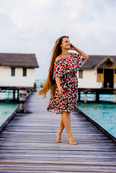 Joyful cute attractive young happy long-hair girl standing on a wooden pier near water bungalows and enjoying ocean, summer breeze and sound of the waves during vacation. Happy holiday travel concept. - Photo, Image