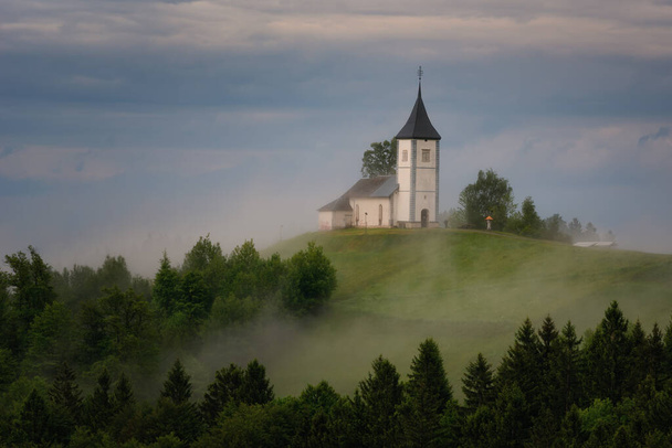 Iglesia Jamnik en una ladera en la primavera, el tiempo brumoso al atardecer en Eslovenia, Europa. Paisaje de montaña poco después de lluvia primaveral. Alpes eslovenos
. - Foto, imagen