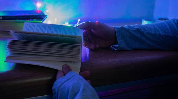 Hands of a white man, holding a book, with three books in the background and colored lights, on a wooden table, with a white background - Photo, image