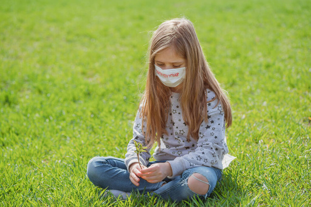 Flu epidemic, self-isolation and coronavirus protection concept.A little girl in a protective mask on her face and text coronavirus is resting on the green grass in nature. - Photo, Image