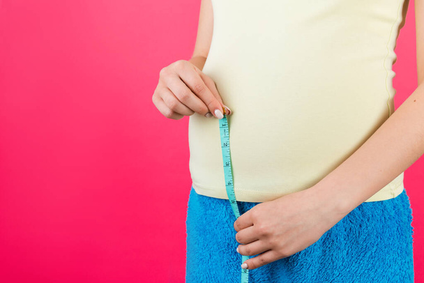 Cropped image of pregnant woman in home clothing measuring her growing abdomen at pink background. Inch tape measure. Copy space. - Photo, Image