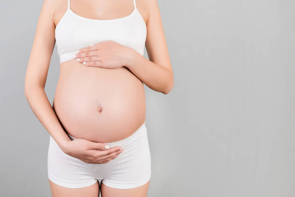 Cropped image of pregnant woman in white underwear holding her belly at gray background. Future mother is waiting her baby. Copy space. - Fotografie, Obrázek