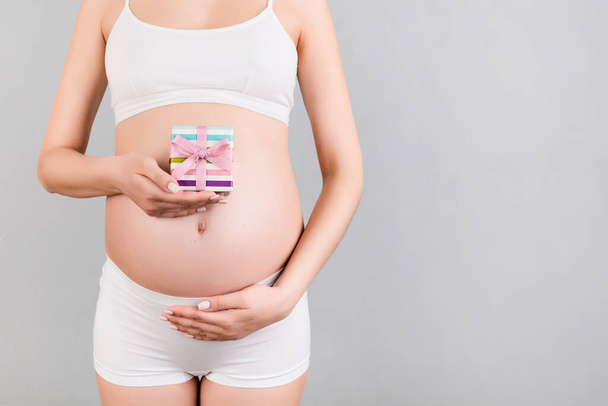 Обрезанное изображение маленькой подарочной коробки в руках беременной женщины на сером фоне. Молодая мать носит белое нижнее белье. Счастливой беременности. Копирование пространства
. - Фото, изображение