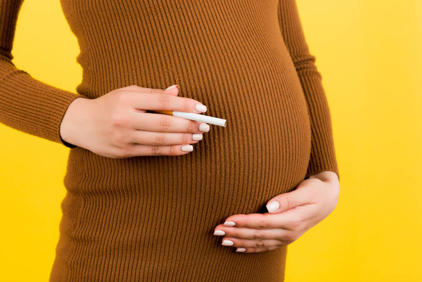 黄色の背景に腹にタバコで茶色の服を着た妊婦のクローズアップ。生まれていない赤ん坊にとって危険なリスク. - 写真・画像