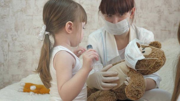 子供たちは医療用注射器で保護マスクをして遊ぶ。ゲームは医者、看護師、ワクチンで患者を治療するふりをします。健康な女の子はおもちゃのクマを注入します。子供は病院で. - 写真・画像
