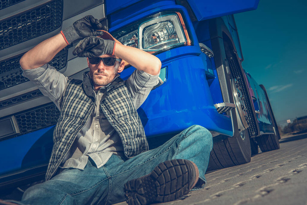 Motorista masculino do caminhão semi fora do veículo que senta-se no solo que relaxa e que limpa a testa na parada do descanso
.  - Foto, Imagem
