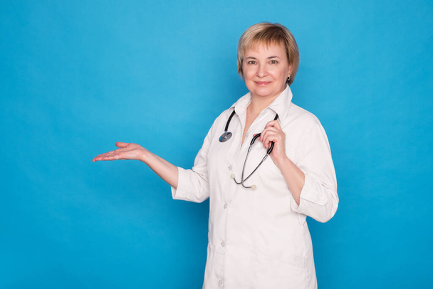 Starší bělošská doktorka v bílém plášti se stetoskopem na modrém pozadí. Emocionální portréty: má něco v ruce. Mezera pro text. - Fotografie, Obrázek