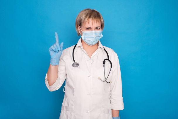 Ältere kaukasische Ärztin in weißem Mantel und mit Stethoskop auf blauem Hintergrund. Emotionale Porträts: Sie zeigt mit dem Finger. - Foto, Bild