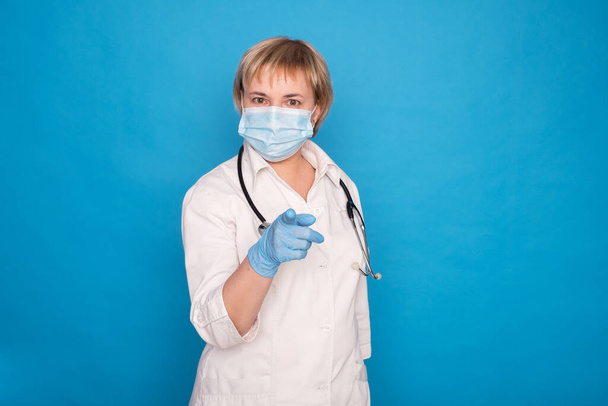 Oudere blanke arts dame in witte jas en met stethoscoop op blauwe achtergrond. Emotionele portretten: ze wijst met haar vingers. - Foto, afbeelding