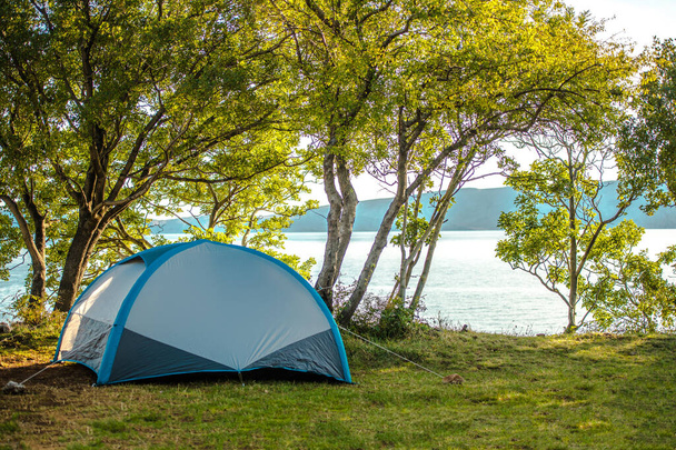 Pohled na malý modrý stan postavený na stínu podle stromů v blízkosti jezera a hor. - Fotografie, Obrázek