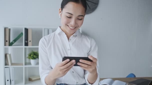 An attractive young asian businesswoman with wireless earphones is watching something on her mobile phone while sitting in the office - Séquence, vidéo