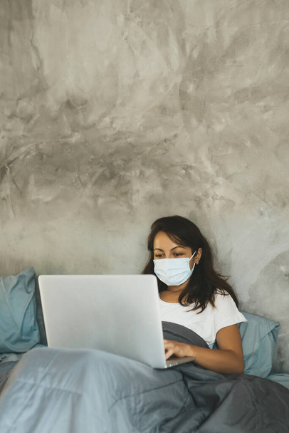 Woman in medical mask laying in bed with laptop. Copy space. - Fotografie, Obrázek