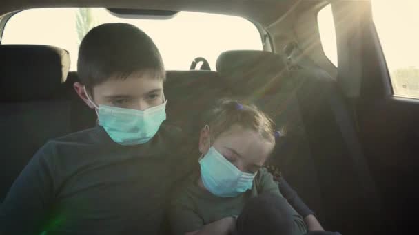 Elokuvatyttö lääketieteellisessä naamiossa nukkuu liikkuvan auton takapenkillä veljensä vieressä. Lasten kasvoilla olevat suojakeinot koronaviruksen ehkäisemiseksi auringonvalossa
 - Materiaali, video