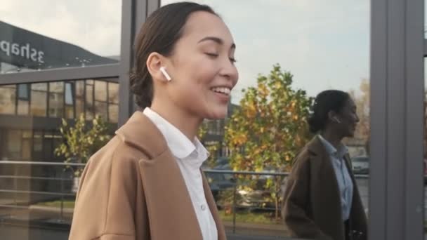 A smiling young asian woman is talking on the phone using her earbuds while walking near modern office buildings - Video