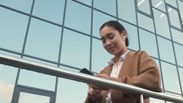 A bottom view of a stylish smiling young asian businesswoman with earbuds is standing near modern office buildings - Filmagem, Vídeo