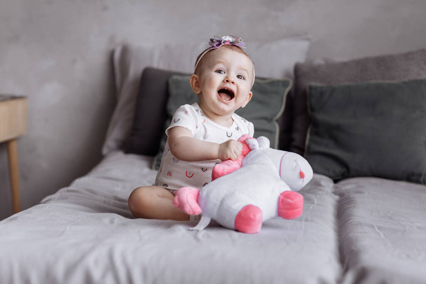 adorable little girl is playing with toy unicorn on bed at home. concept of childhood day. happy baby's, family day. - 写真・画像
