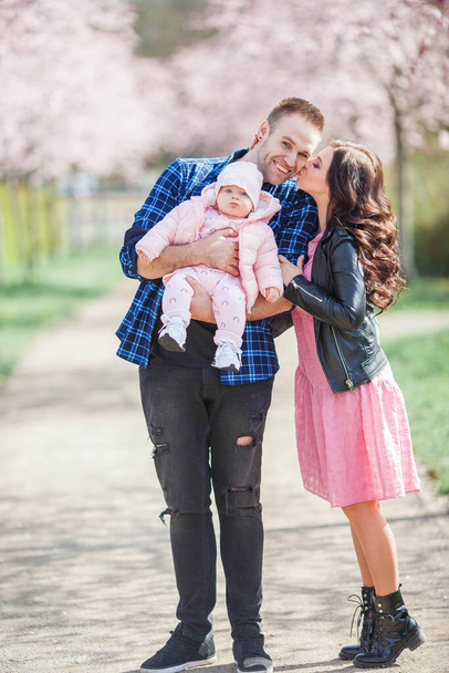 A fiatal szülők egy csinos kislányt tartanak a karjaikban. Gyönyörű család egy virágos kertben. Sakura vagyok. Tavasz. - Fotó, kép