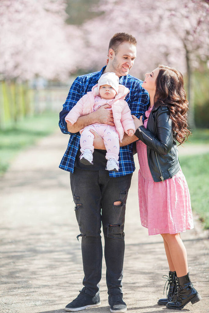 Jonge ouders houden een kleine mooie dochter in hun armen. Mooie familie in een bloemrijke tuin. Sakura. Voorjaar. - Foto, afbeelding