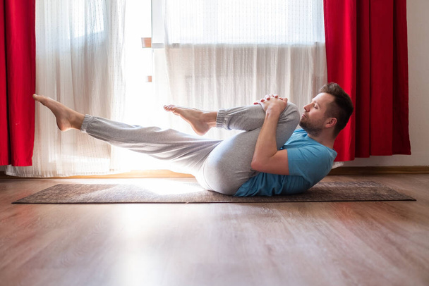 Kafkasyalı adam yerde yoga yapıyor, desteklenen tek bacaklı rüzgar dindirme pozu üzerinde çalışıyor ya da Eka Pada Pavanamuktasana evde oturma odasında. - Fotoğraf, Görsel