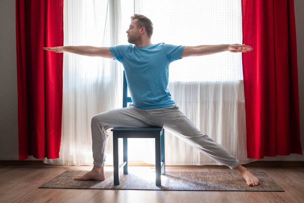 Caucasian young man in Warrior 2 Pose using chair at his living room at home - Photo, Image