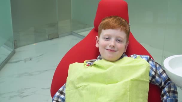 Gelukkige roodharige jongen bij de receptie bij tandarts in tandartsstoel dat is naar beneden. Pediatrische tandheelkunde - Video