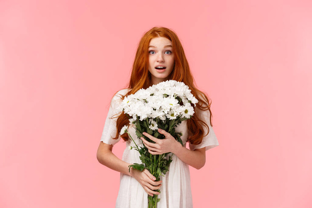 Birthday, celebration and valentines day concept. Surprised romantic and alluring redhead lovely girlfriend receive gift of beautiful white flowers, embracing bouquet and looking touched - Photo, Image