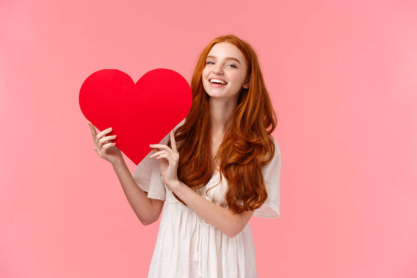 Geluk, liefde en relatie concept. Mooi en schattig roodharig tienermeisje lachen en glimlachen camera, met grote rode hart vrolijk uiten sympathie, diepe gevoelens, roze achtergrond - Foto, afbeelding