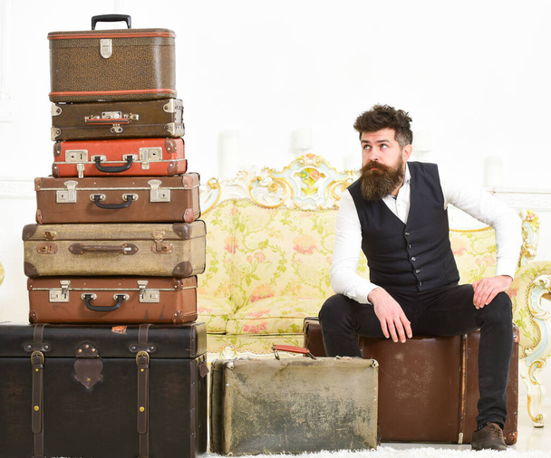 Luggage and relocation concept. Man with beard and mustache packed luggage, white interior background. Macho elegant on tired face sits, exhausted at end of packing, near pile of vintage suitcases. - Fotografie, Obrázek