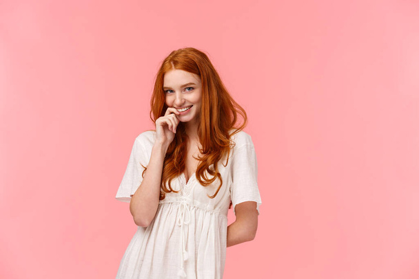 Romance, relationship and tenderness concept. Beautiful tender redhead woman in cute white dress, blushing and giggle silly, smiling biting finger gazing at camera sensual, flirt over pink background - Photo, Image