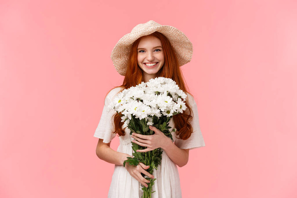 Ρομαντικό δελεαστικό υπέροχο κορίτσι κοκκινομάλλα πρόκειται για μια βόλτα, απολαμβάνοντας την άνοιξη, φορώντας καπέλο ήλιο και κρατώντας λουλούδια μπουκέτο ως έχοντας τέλεια ημερομηνία με το φίλο, στέκεται ροζ φόντο - Φωτογραφία, εικόνα