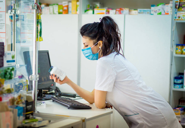 the pharmacist is holding a container with medicine and is studying it. A pharmacist in a medical mask on his face punches medicine in a computer. - Photo, Image
