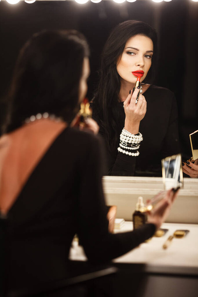 Jewelry and beauty concept. Beautiful brunette lady portrait in modern gems and black dress in studio. Woman sits by the makeup mirror and puts on red lipstick - Foto, imagen