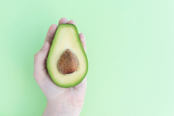 hand holds half of avocado with pit on light green background, space for text, healthy food concept, dieting and detoxing nutrition - 写真・画像