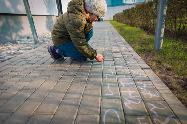 pieni tyttö kirjoittaa numeroita asfalttiin, lapsi leikkii ulkona, kun koulu on suljettu
 - Valokuva, kuva