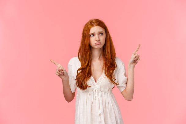 Waist-up portrait innocent, cute and timid redhead indecisive girl, dont know what do, pouting feeling sad, gloomy look around, pointing fingers left and right, pink background - Foto, Imagen