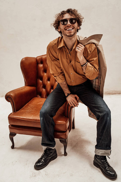 Retrato de un chico casual guapo con el pelo rizado posando en el estudio brillante mientras está sentado en la silla de cuero
 - Foto, imagen
