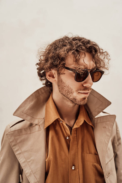 Bedachtzame en krullende man draagt een zonnebril poseren in de heldere studio op zoek rijk - Foto, afbeelding