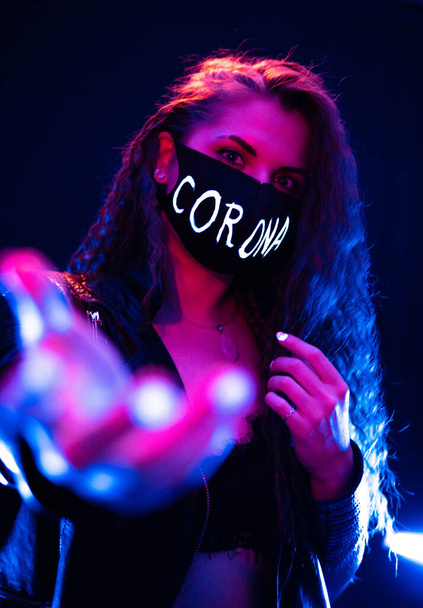Πορτρέτο ενός Beckoning ελκυστικό κορίτσι φορώντας μαύρη προστατευτική μάσκα με στέμμα Είσοδος στο πρόσωπο σε νέον φώτα με μαύρο ομιχλώδες φόντο - Φωτογραφία, εικόνα